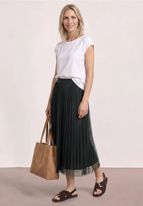 Frau in weißem T-Shirt, schwarzem Plisseerock, schwarzen Sandalen und großer brauner Tasche.
