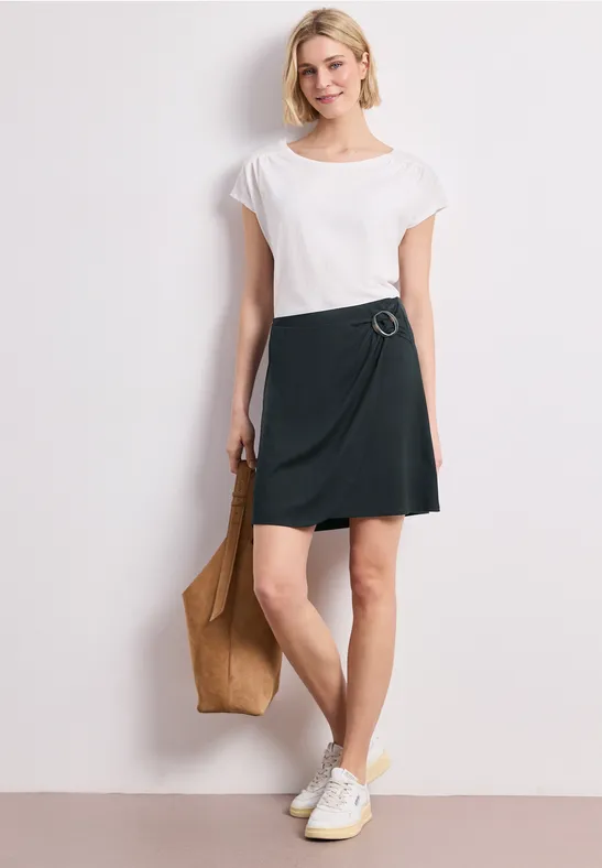 Eine Frau in weißem T-Shirt, dunkelgrünem Wickelrock, weißen Sneakern und brauner Tragetasche.