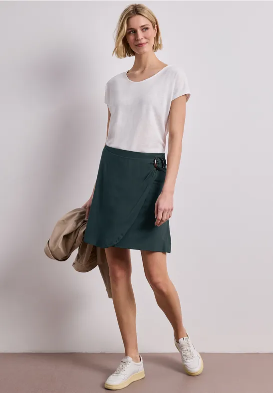 Frau trägt weißes T-Shirt mit grünem Wickelrock und Sneakers, hält eine Jacke in der Hand.