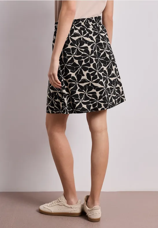 Schwarzer A-Linien-Rock mit weißem Blumenmuster und beigen Espadrilles vor hellem Hintergrund.