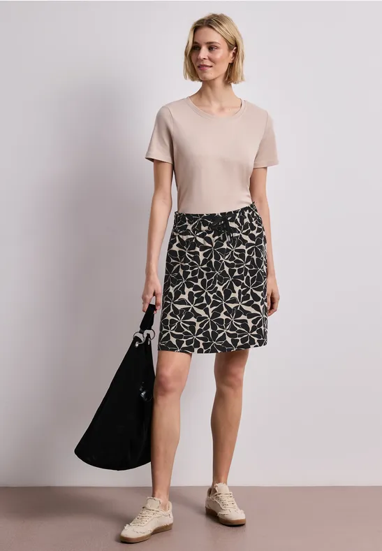 Frau in beigem T-Shirt, gemustertem Rock, weißen Sneakers und schwarzer Tasche vor neutralem Hintergrund.