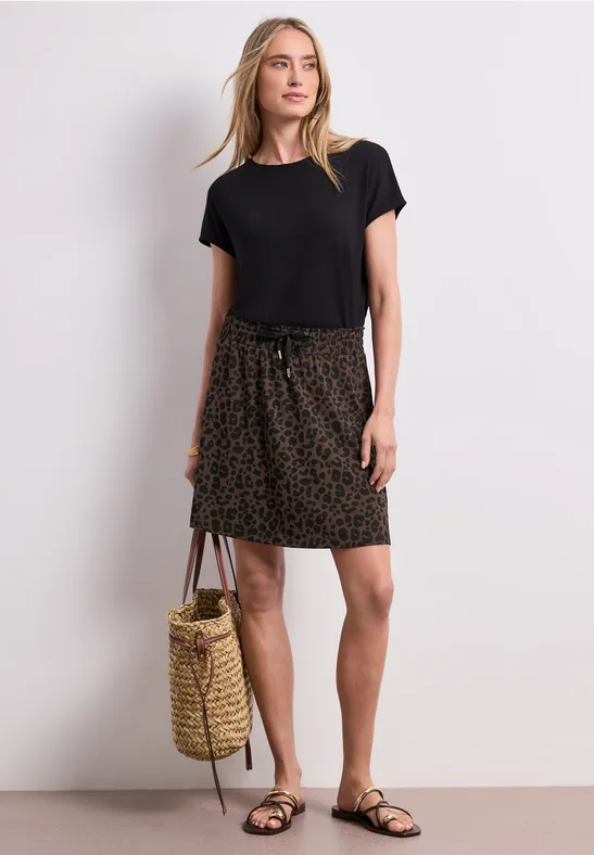 Frau in schwarzem Shirt, leopardgemustertem Rock und Sandalen, hält eine Korbtasche vor weißem Hintergrund.