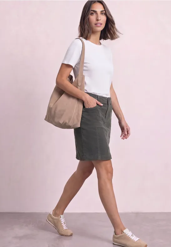 Vrouw in wit t-shirt, groene rok en sneakers, met een beige tas voor een roze achtergrond.