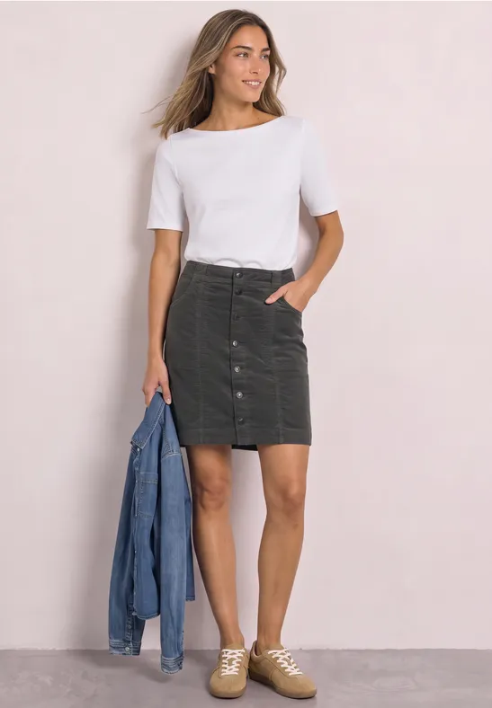 Vrouw in witte T-shirt, groene corduroy minirok en sneakers, houdt spijkerjack vast