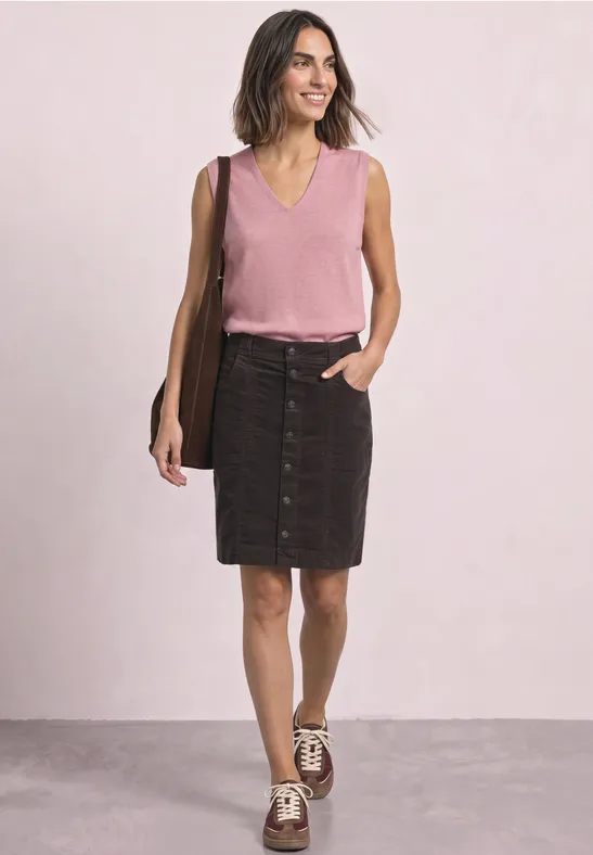 Vrouw in roze mouwloos topje en zwarte jeansrok met knopen, bruin schoudertas en sneakers voor lichte achtergrond.
