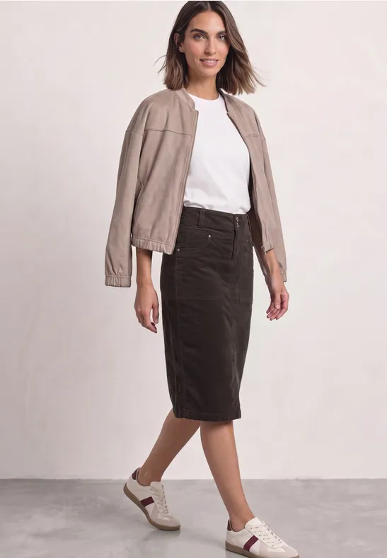 Une femme porte un t-shirt blanc, une veste en cuir beige, une jupe crayon noire et des baskets blanches.