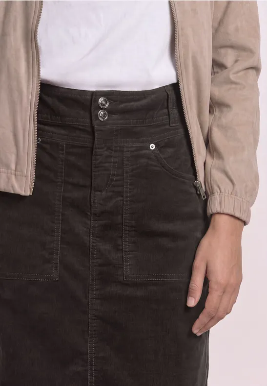Une personne porte un t-shirt blanc, une veste beige et une jupe en velours côtelé noire avec poches.