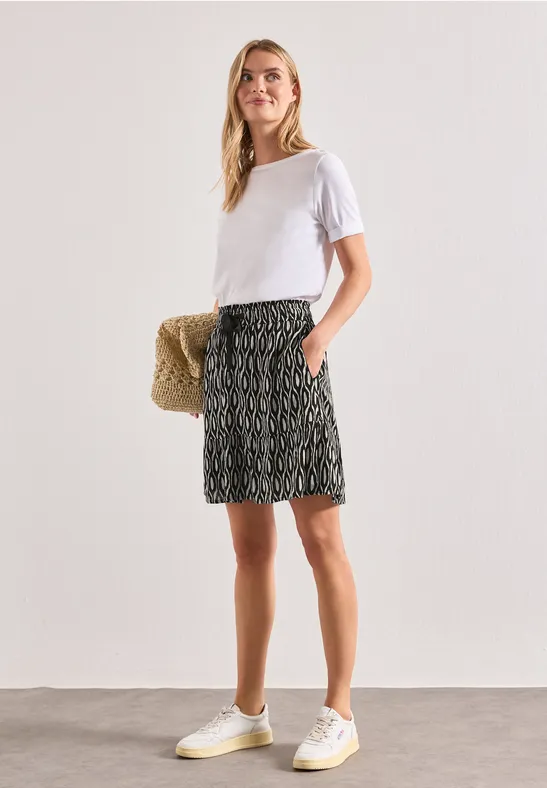 Vrouw in wit T-shirt, rok met dessin en witte sneakers, met een strooien tas.