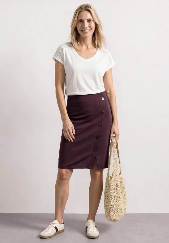 Eine Frau in weißem T-Shirt, bordeauxfarbener Knopfrock, weiße Sneaker und netzartige Tasche