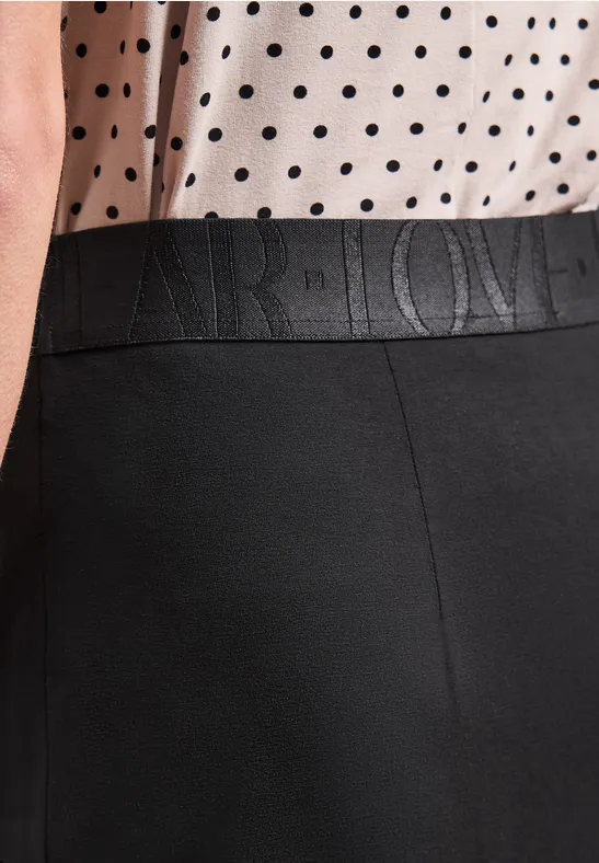 Vrouw draagt wit topje met zwarte stippen en een zwarte broek met geperst logo in de taille.