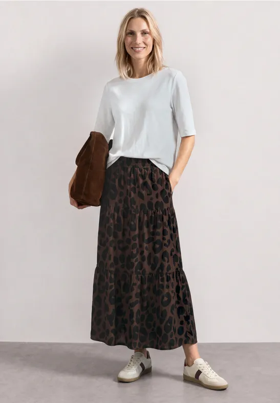 Frau in weißem T-Shirt und langem Leoparden-Rock, Sneakern und brauner Jacke.