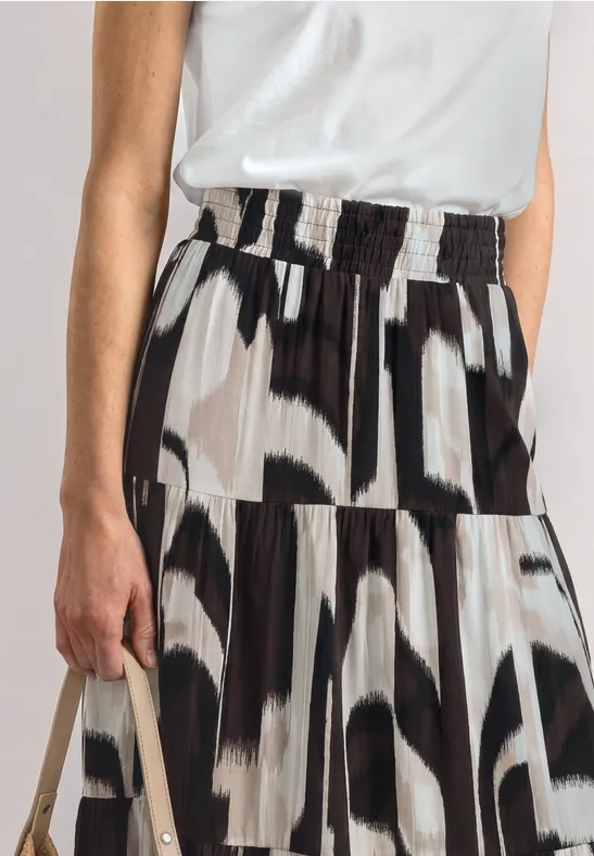Persoon draagt wit topje en geplooide rok met abstract zwart-wit patroon en houdt een beige tas vast.