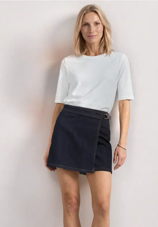 Model trägt weißes T-Shirt und dunklen Jeans-Minirock vor hellem Hintergrund.