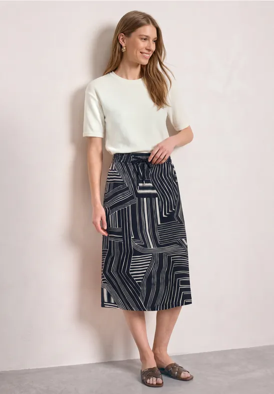 Frau vor hellem Hintergrund mit weißem T-Shirt, schwarzem Rock mit geometrischem Muster und schwarzen Sandalen.