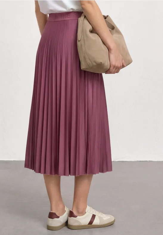 Une personne porte une jupe midi plissée violette, des baskets blanches à détails rouges et un sac beige.