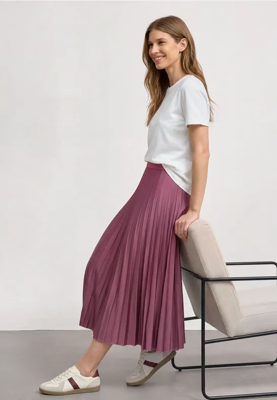 Une femme porte un t-shirt blanc et une jupe plissée rose, assise de profil sur une chaise.