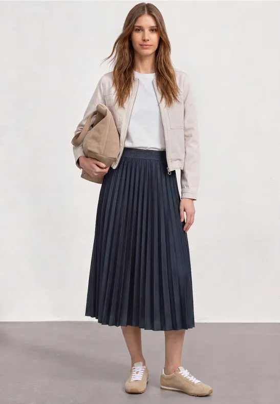Frau mit beigem Wildlederblazer, weißem Shirt, knielangem dunkelblauem Faltenrock und beigen Sneakern hält Clutch.