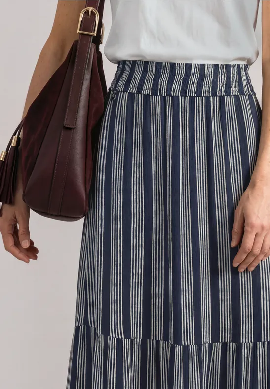 Femme en haut blanc et jupe longue rayée bleu et blanc, avec un sac marron à l’épaule.