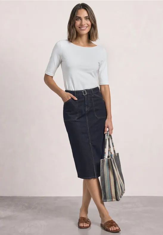 Frau in weißem Oberteil, dunklem Jeans-Midirock und Sandalen hält gestreifte Tasche.
