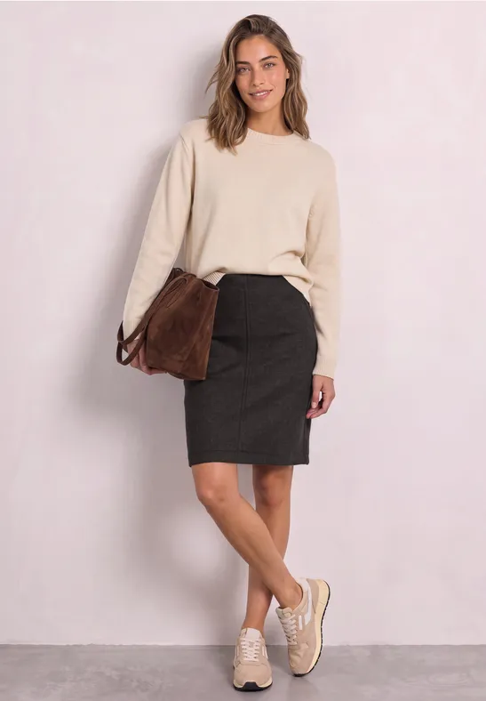 Junge Frau trägt cremefarbenen Pullover, schwarzen Bleistiftrock, Sneaker und eine Tasche.