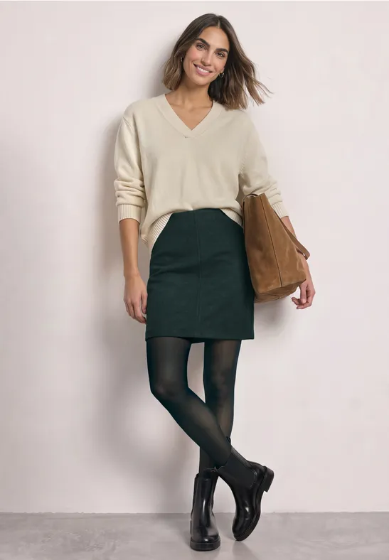 Eine Frau in beigem V-Pullover, schwarzem Rock, schwarzen Strumpfhosen und Stiefeletten mit brauner Tasche.