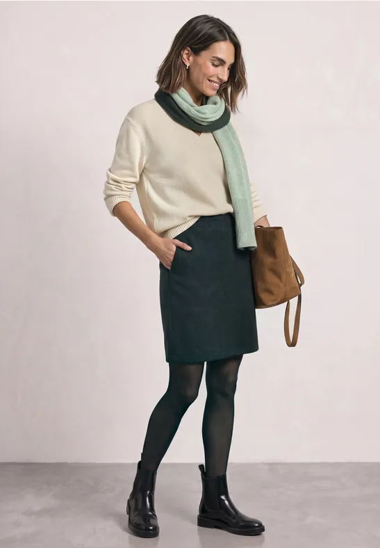 Frau in beigem Pullover, grünem Schal, dunklem Rock, schwarzen Strumpfhosen und Boots mit brauner Tasche.
