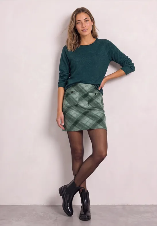 Frau mit grünem Pullover, karriertem Minirock, schwarzen Strumpfhosen und Boots vor hellem Hintergrund.