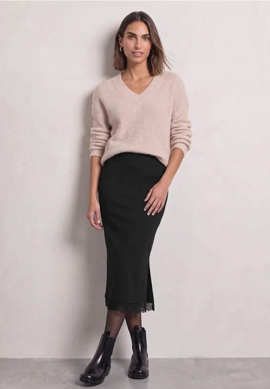 Vrouw in beige V-hals trui, zwarte midi rok met kant en enkellaarsjes voor een lichte achtergrond.