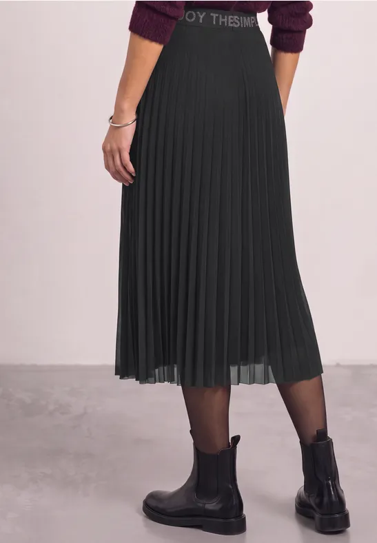 Zijaanzicht van een persoon in een zwarte, geplooide midirok met zwarte panty's en enkellaarsjes.