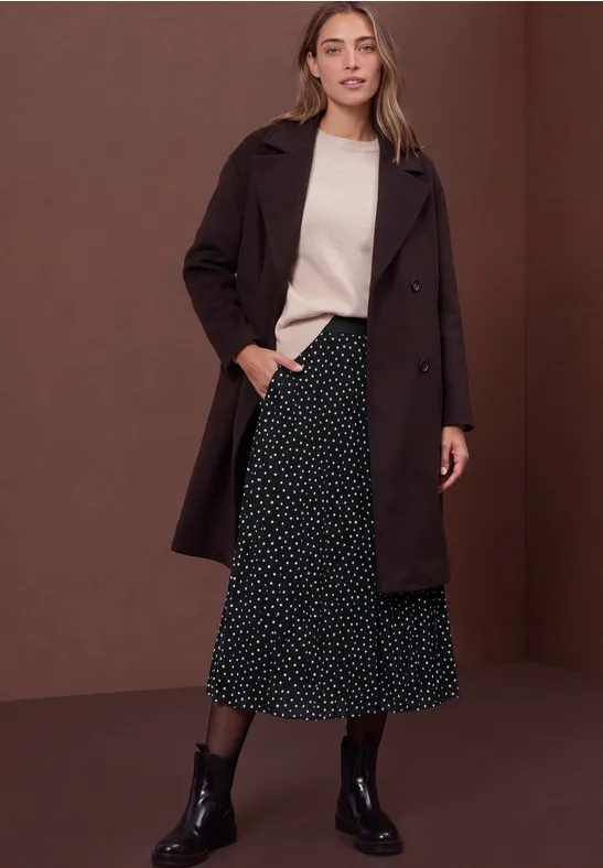 Frau mit dunkelbraunem Mantel, hellem Pullover, schwarzem gepunktetem Midirock und schwarzen Stiefeln vor braunem Hintergrund.