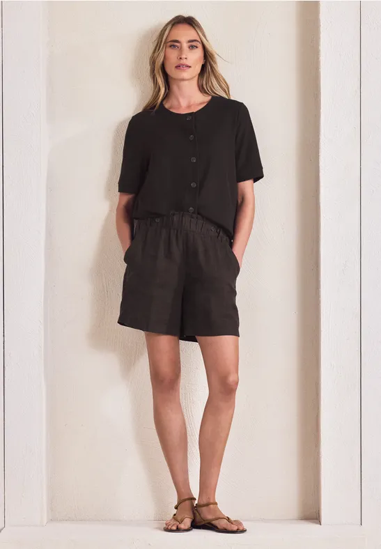 Frau in schwarzem T-Shirt, kurzen Hosen und Sandalen steht vor einer hellen, schlichten Nische.