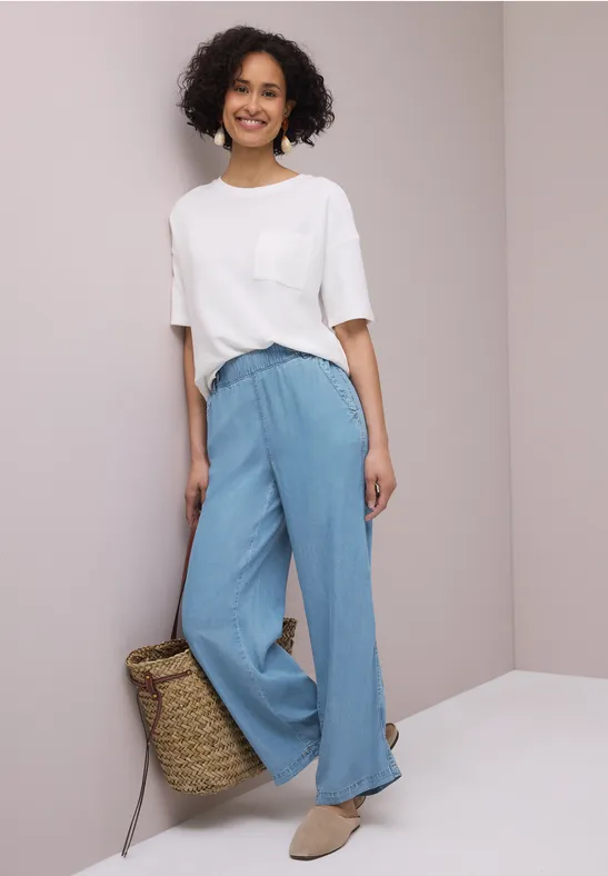 Vrouw in wit t-shirt, losse lichtblauwe broek en strooien tas, voor een lichte achtergrond.