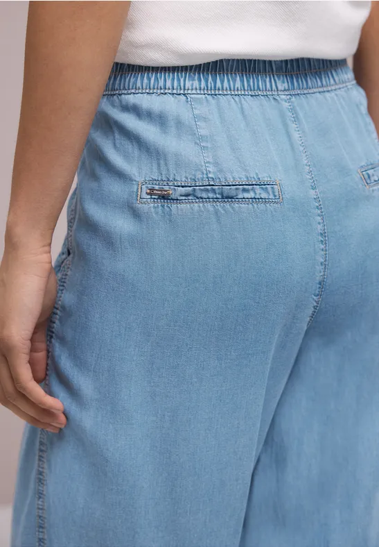 Achteraanzicht van een persoon in lichtblauwe, losse denimbroek met elastische tailleband, hand langs het lichaam.