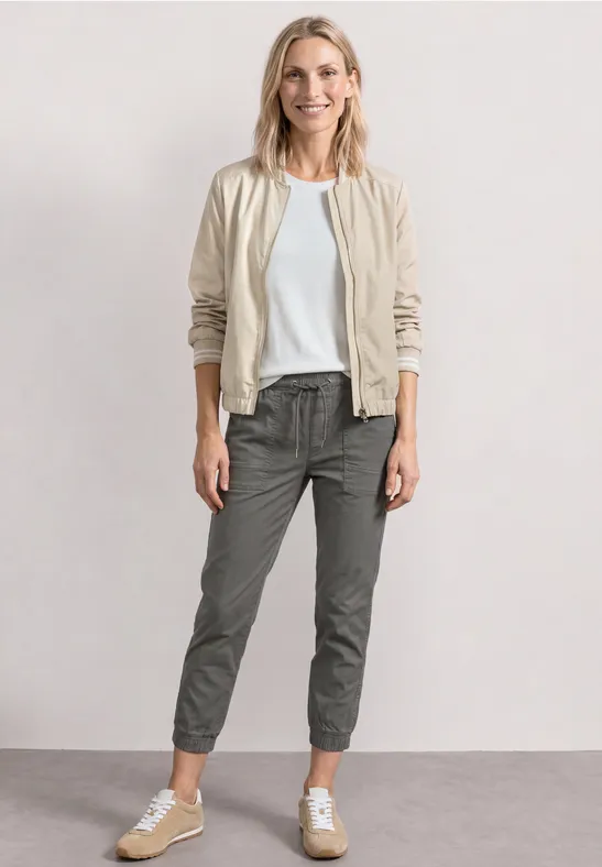 Une femme en pose décontractée porte une veste bomber beige, un t-shirt blanc, un pantalon gris court et des baskets.
