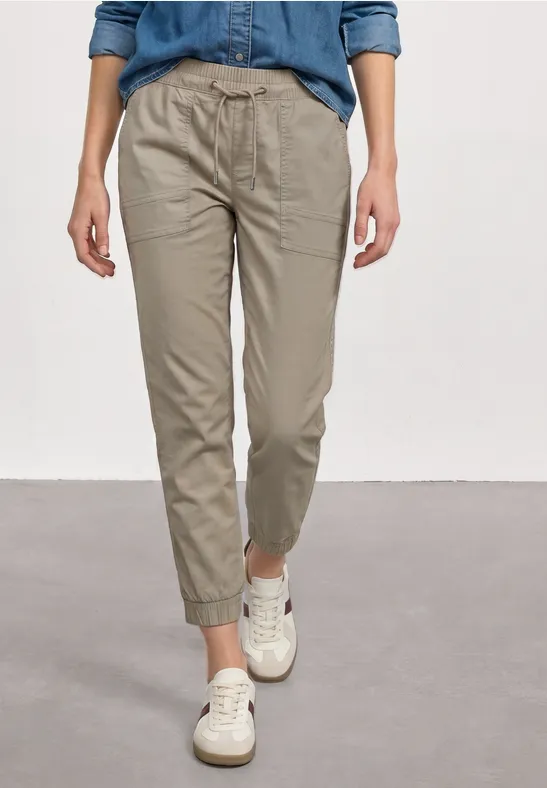 Persoon in beige chino met elastische taille, opgerolde pijpen en witte sneakers voor een grijze achtergrond