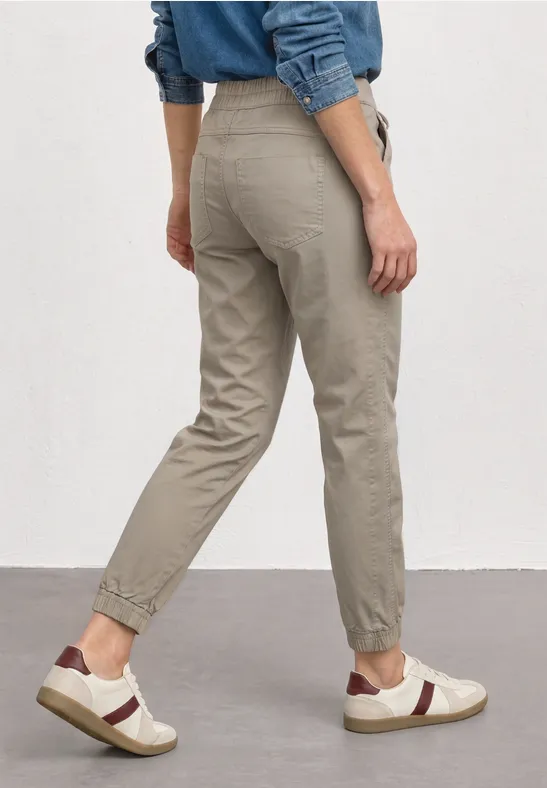 Persoon van achteren in beige joggingsbroek en witte sneakers, blauwe spijkershirt tegen witte muur.