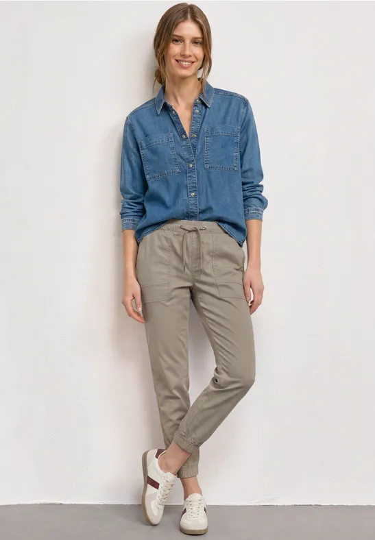 Vrouw in jeansoverhemd, beige chino en witte sneakers tegen lichte achtergrond.