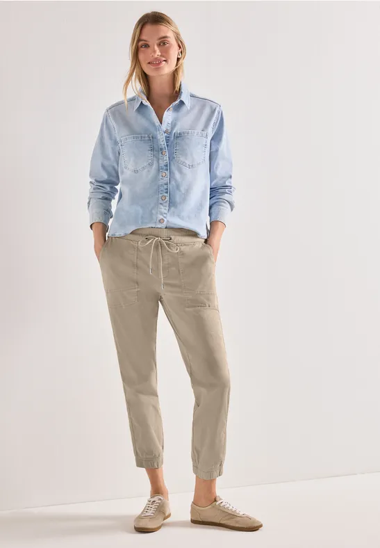 Vrouw in lichtblauw spijkerblouse, beige joggingbroek en sneakers, staand voor een lichte, neutrale achtergrond.