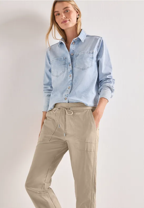 Vrouw in lichtblauw denim blouse en beige, losse broek met trekkoord en steekzakken tegen witte achtergrond.