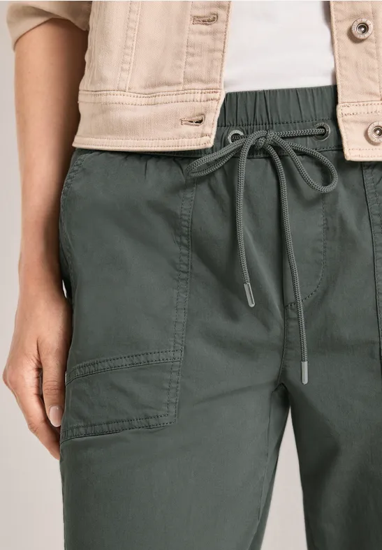 Close-up van een donkergroene broek met elastische taille en koord, met een licht jasje erboven.