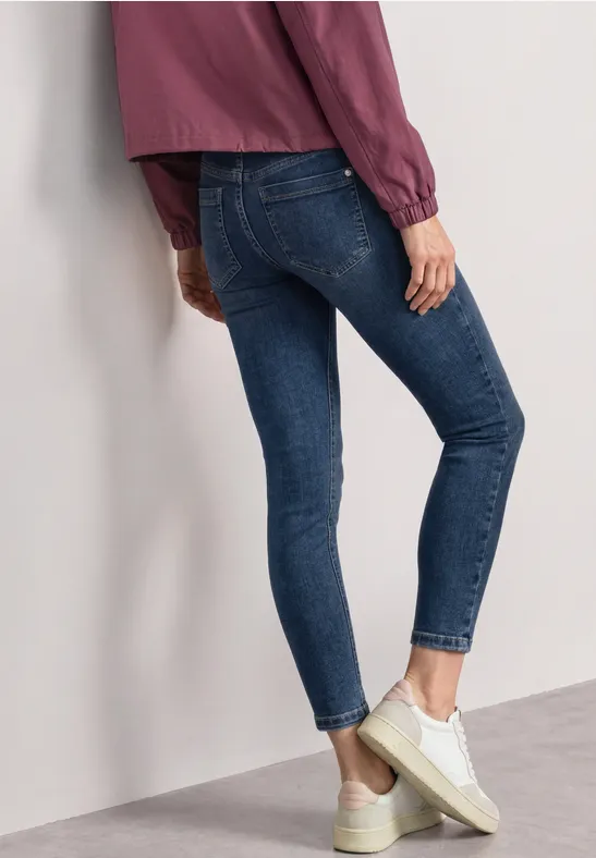 Person in engen blauen Jeans, weißem Sneaker und lila Oberteil, seitlich an Wand gelehnt.