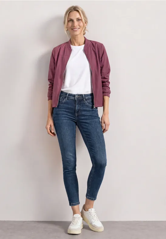 Frau in weißem T-Shirt, bordeauxfarbener Jacke, blauen Skinny Jeans und weißen Sneakern vor hellem Hintergrund.