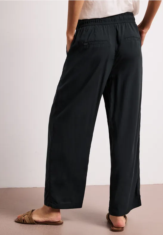 Achteraanzicht van een persoon met zwarte, wijde broek met elastische taille en bruine sandalen