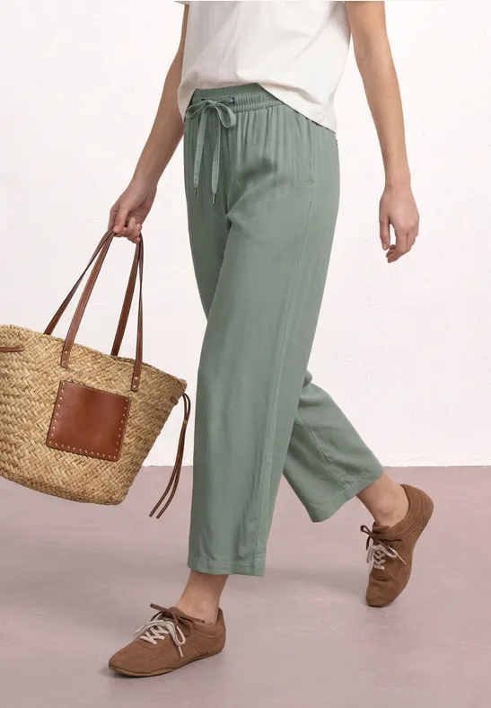 Person trägt grüne, weiche Hose, weißes T-Shirt, braune Schnürschuhe und eine Strohtasche.