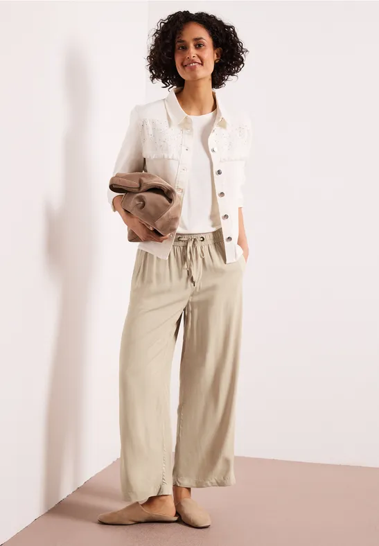 Een vrouw draagt wijde beige linnen broek, een witte jas en houdt een bruine jas vast.