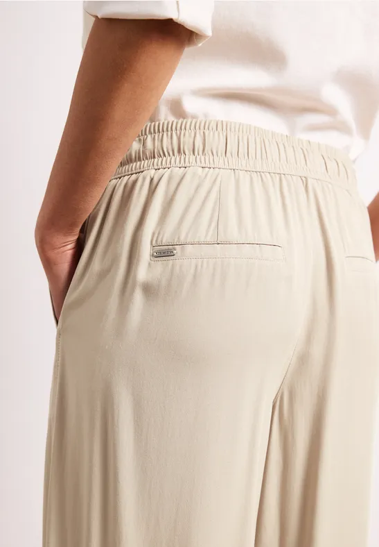 Een persoon draagt een beige losse broek met elastische taille en achterzak met rits, hand in de zijzak.