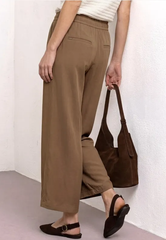 Vrouw van achteren in losse, bruine broek, witte top, zwarte sandalen en een bruine schoudertas.