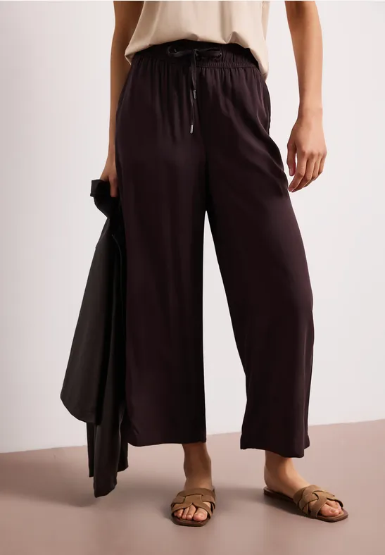 Losse, zwarte broek met trekkoord, gecombineerd met bruine sandalen en een bijpassend jasje in de hand.