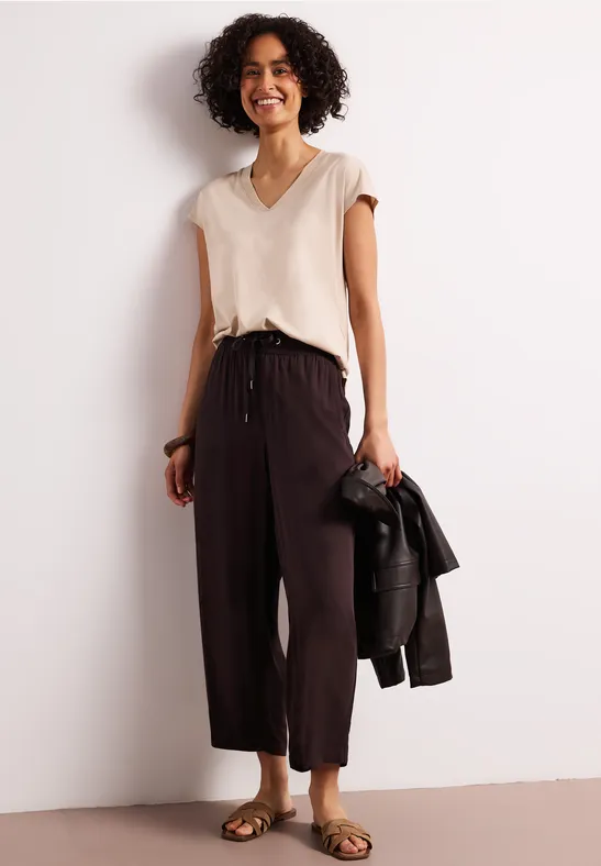 Vrouw met krullend haar draagt een beige top en zwarte wijdvallende broek, houdt een zwarte leren jas vast.