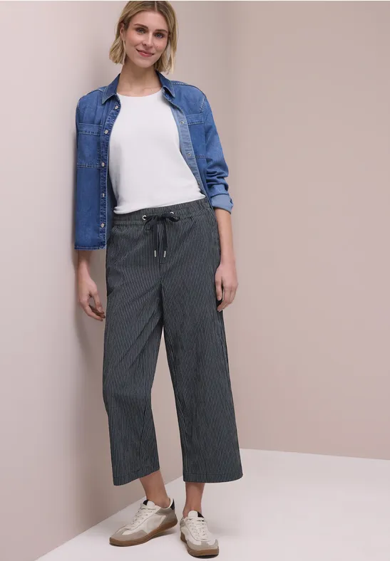 Een persoon draagt een grijze broek, wit t-shirt, spijkerjasje en witte sneakers voor een beige muur.